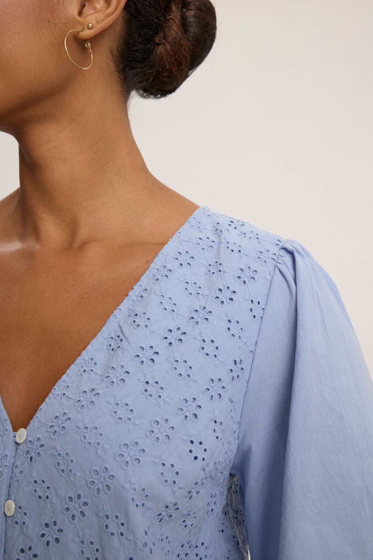 A woman wearing the NELLI EYELET DRESS by Velvet by Graham & Spencer, a light blue embroidered cotton dress with a V neckline, and hoop earrings, shown from the shoulders up against a plain background.-39123388989633