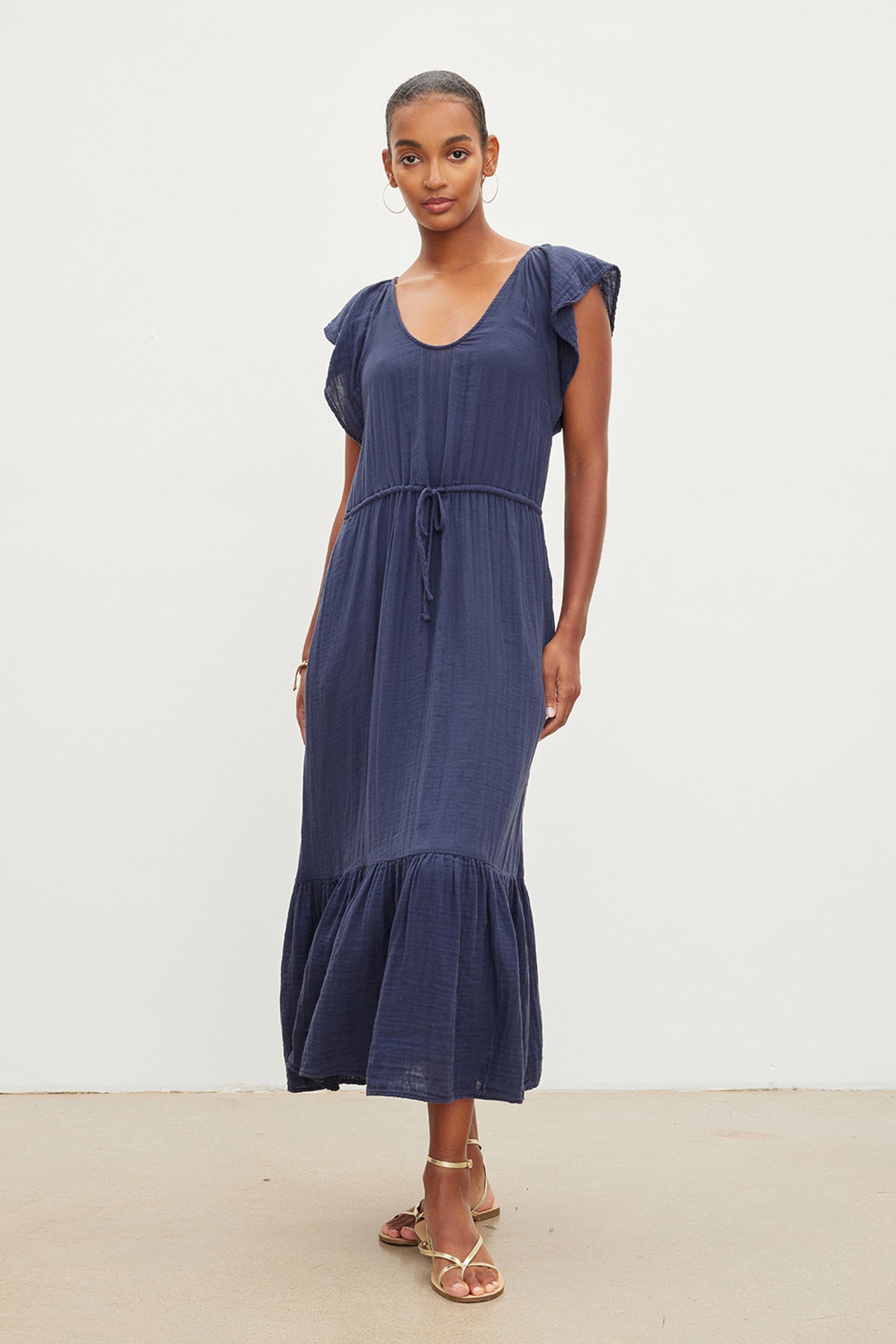 A woman in a navy blue scoop neckline dress with ruffled sleeves and a tiered skirt, wearing gold sandals, stands confidently against a plain white background in the Velvet by Graham & Spencer ADALYN COTTON GAUZE TIERED DRESS.