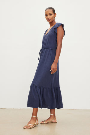 A woman in a navy scoop neckline dress with drawstring waist and ruffle sleeves, paired with gold sandals, standing against a plain background, wearing the ADALYN COTTON GAUZE TIERED DRESS by Velvet by Graham & Spencer.