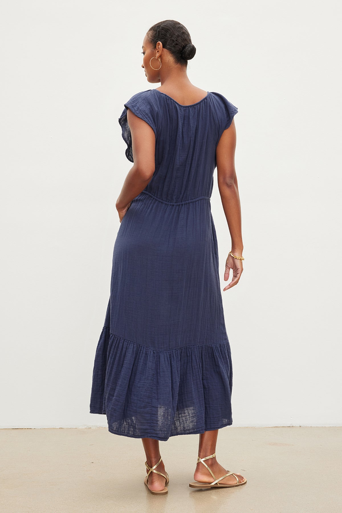 A woman viewed from behind, wearing a Velvet by Graham & Spencer ADALYN COTTON GAUZE TIERED DRESS and gold sandals, standing against a neutral background.