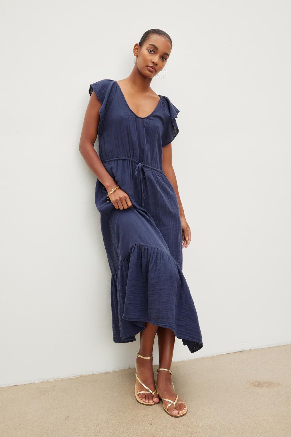 A woman posing in a navy blue scoop neckline dress with cap sleeves and a tie waist, paired with rose gold sandals, standing against a neutral background. The ADALYN COTTON GAUZE TIERED DRESS by Velvet by Graham & Spencer.-36443668414657