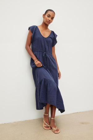 A woman posing in a navy blue scoop neckline dress with cap sleeves and a tie waist, paired with rose gold sandals, standing against a neutral background. The ADALYN COTTON GAUZE TIERED DRESS by Velvet by Graham & Spencer.