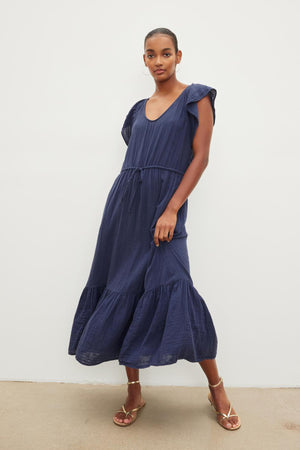 A woman stands in a studio, wearing a dark blue ADALYN COTTON GAUZE TIERED DRESS from Velvet by Graham & Spencer with ruffle sleeves and gold sandals, looking at the camera. The dress features a scoop neckline.