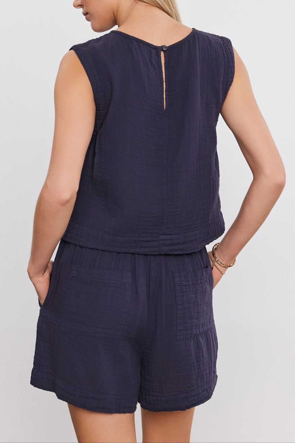 Woman wearing the AUBREN COTTON GAUZE TANK TOP by Velvet by Graham & Spencer, a sleeveless, navy blue top and matching shorts, viewed from the back. Made of airy cotton gauze, the ensemble features a cropped silhouette that adds to its casual charm. She has her hands in her pockets.