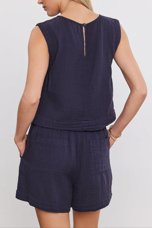 Woman wearing the AUBREN COTTON GAUZE TANK TOP by Velvet by Graham & Spencer, a sleeveless, navy blue top and matching shorts, viewed from the back. Made of airy cotton gauze, the ensemble features a cropped silhouette that adds to its casual charm. She has her hands in her pockets.