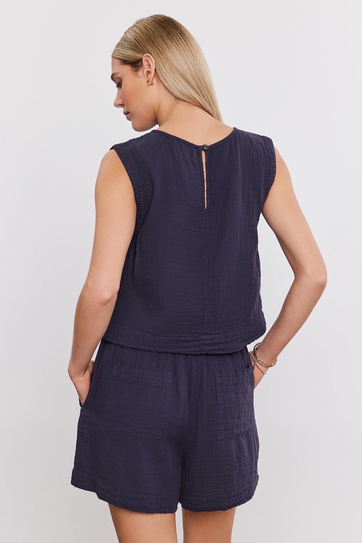Woman with blonde hair wearing a navy sleeveless top and matching TIBBY COTTON GAUZE SHORT by Velvet by Graham & Spencer with a drawstring elasticized waist, standing with hands in pockets, facing away.