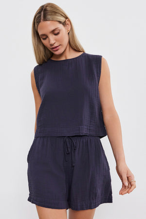 A woman with long, straight blonde hair is wearing a sleeveless, dark blue top and matching Velvet by Graham & Spencer TIBBY COTTON GAUZE SHORT. She is looking down with one hand slightly raised.