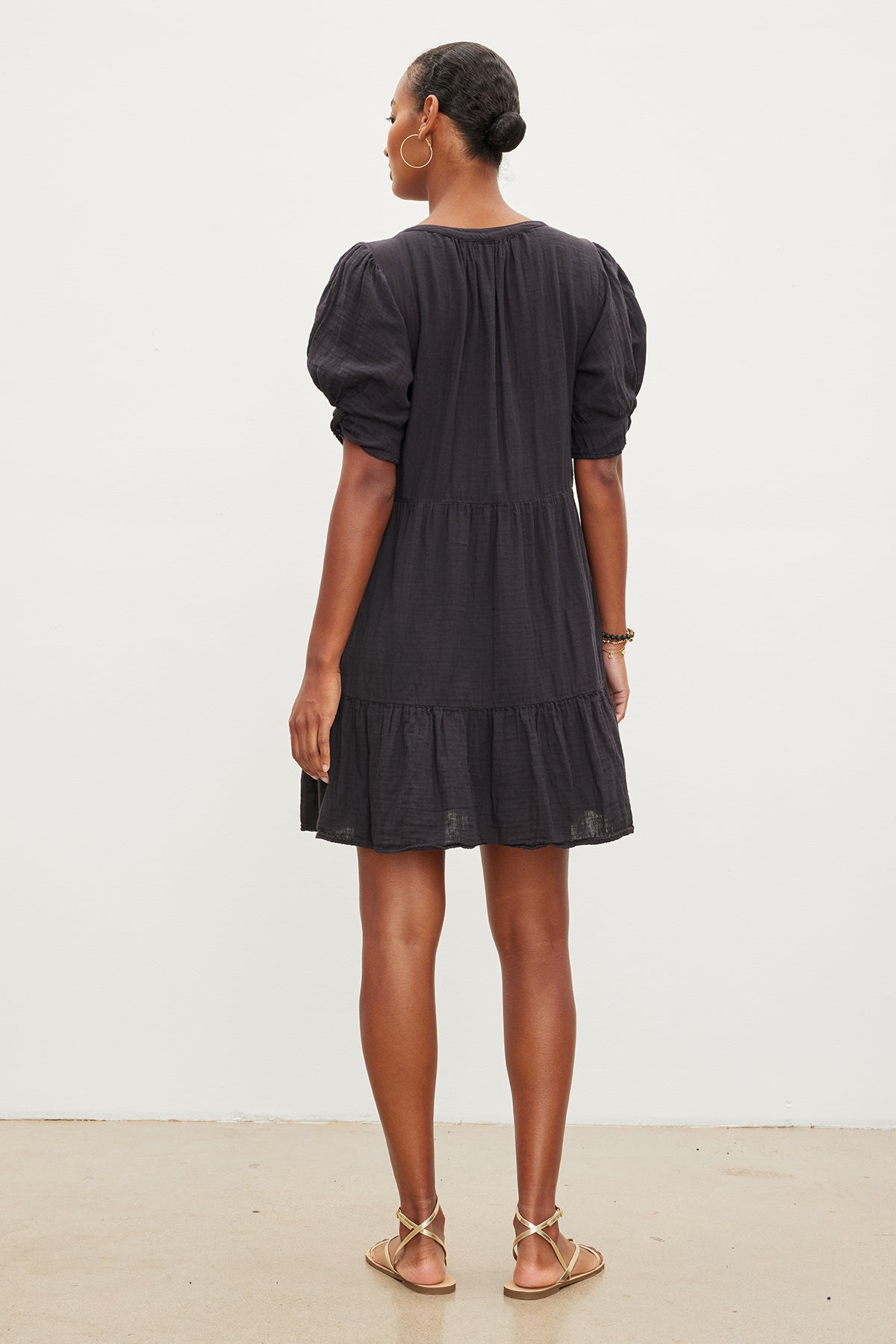 A woman stands facing away, wearing a dark grey BELLA COTTON GAUZE DRESS with puff sleeves and gold sandals by Velvet by Graham & Spencer.-35955428294849