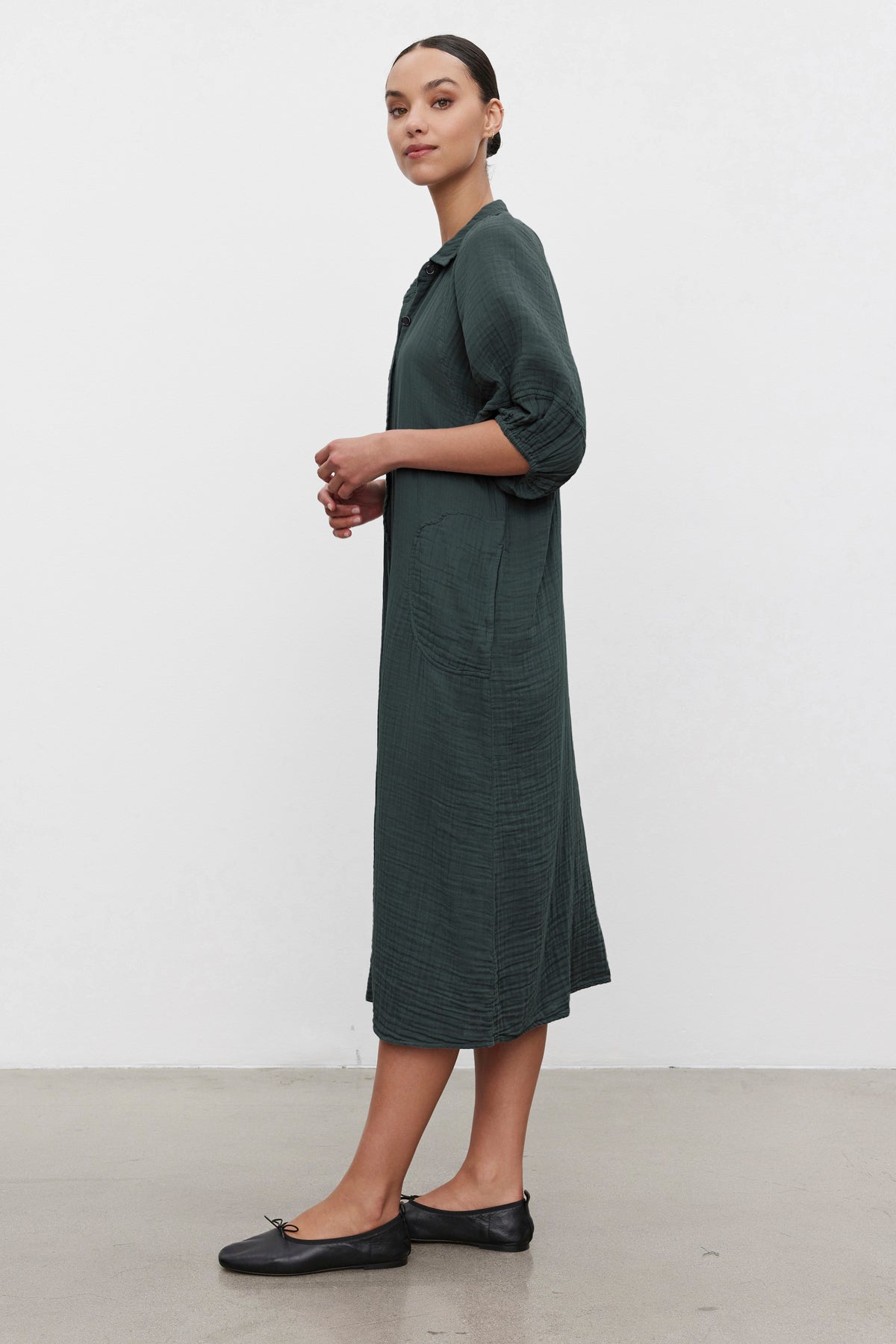 A person stands in profile against a plain background, wearing the BREANA COTTON GAUZE DRESS from Velvet by Graham & Spencer— it's a long dark green button-front dress with rolled-up sleeves paired with black flats.-37676277235905