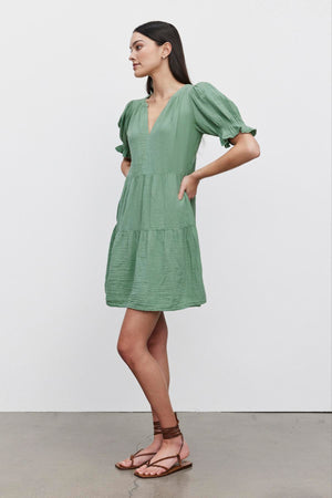 A woman standing in a Velvet by Graham & Spencer CLARISSA COTTON GAUZE TIERED DRESS with puffed sleeves and tiered skirt, paired with brown strappy sandals, against a white background.