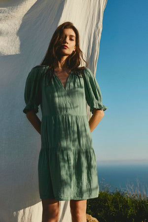 A woman in a Velvet by Graham & Spencer CLARISSA COTTON GAUZE TIERED DRESS with puffed sleeves stands beside a flowing white fabric with a sea background under clear skies.