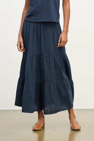 A person stands against a plain backdrop, wearing the DANIELLE COTTON GAUZE SKIRT by Velvet by Graham & Spencer and brown flats.