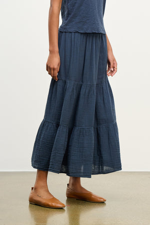A person elegantly poses on a polished floor in the DANIELLE COTTON GAUZE SKIRT by Velvet by Graham & Spencer, featuring a blue tiered midi-to-maxi design with an elastic waist, complemented by brown flats.