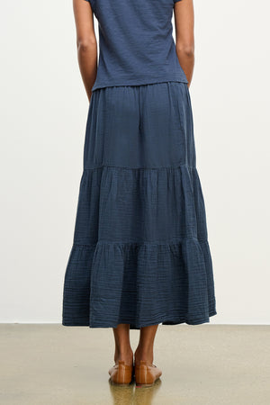 Someone in a DANIELLE COTTON GAUZE SKIRT by Velvet by Graham & Spencer, characterized by its navy tiers and elastic waist, pairs it with brown shoes while standing on a plain floor, captured from the back.