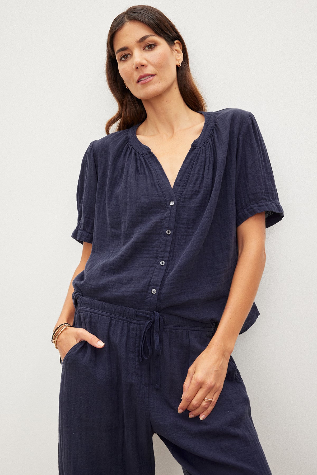 A woman in a navy blue DEANN COTTON GAUZE TOP by Velvet by Graham & Spencer with a button front and matching pants stands with her hand in her pocket against a plain background.-35955697516737