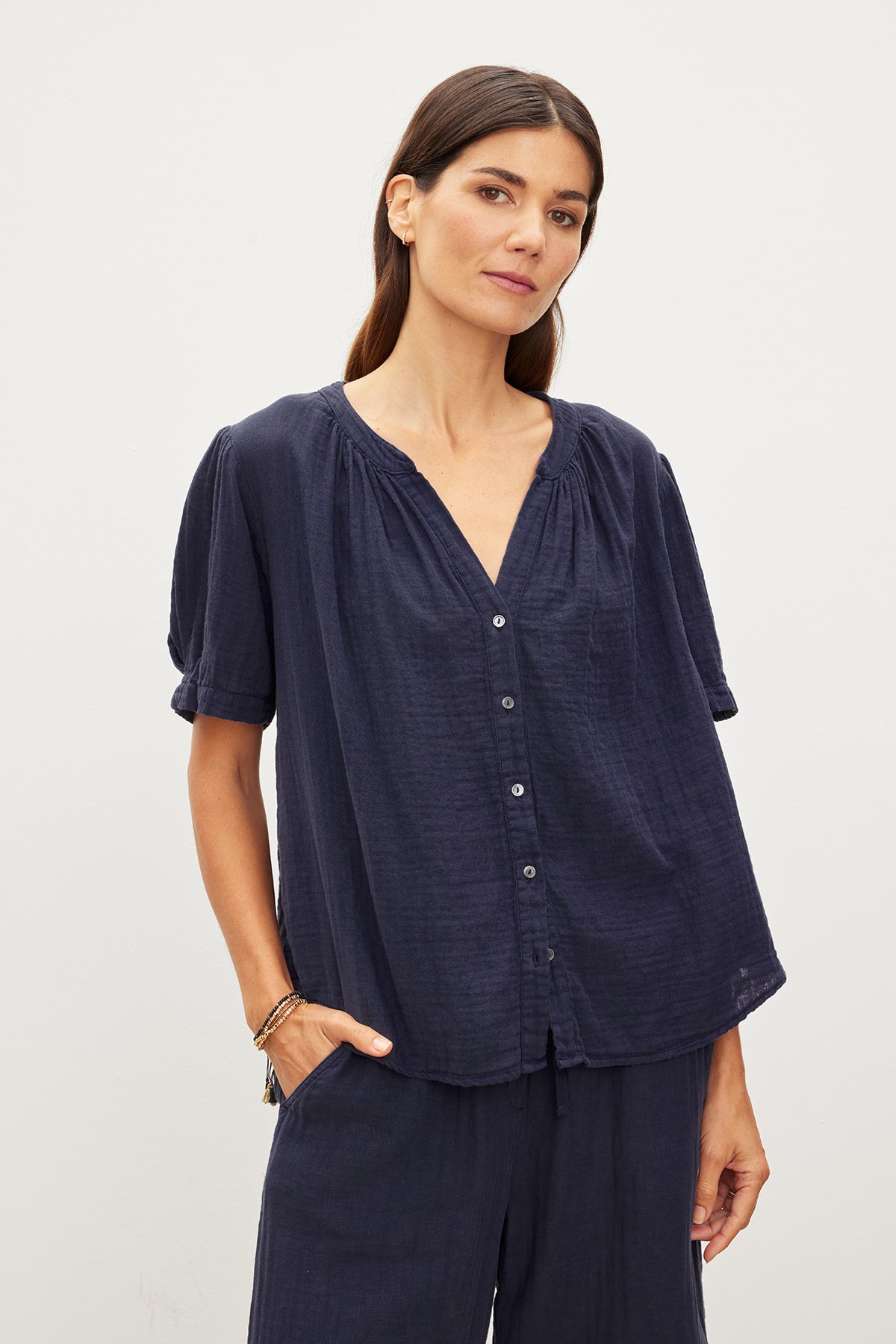 A woman with long dark hair wearing a Navy Blue DEANN COTTON GAUZE TOP by Velvet by Graham & Spencer and matching pants stands against a plain white background. She has one hand by her side and the other in her pocket.