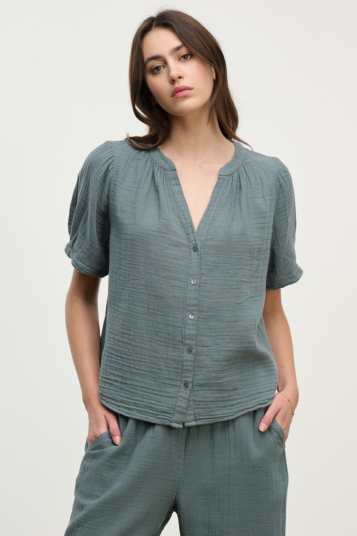 A person is wearing the DEANN COTTON GAUZE TOP by Velvet by Graham & Spencer, a casual green button-up with a subtle v-neckline, and matching pants, hands in pockets against a plain background.