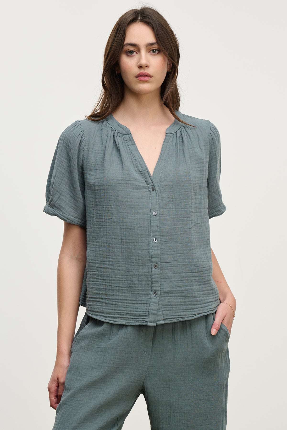 A woman wearing the DEANN COTTON GAUZE TOP, a matching green button-up with a v-neckline crafted by Velvet by Graham & Spencer, paired with pants made from soft cotton gauze, stands with one hand in her pocket.
