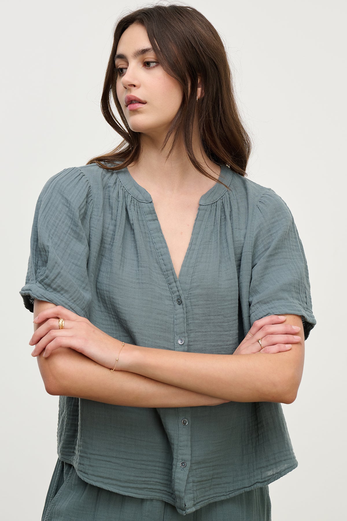 A person with long hair wears Velvet by Graham & Spencer's DEANN COTTON GAUZE TOP, a loose-fitting green button-up with a v-neckline, arms crossed and glancing sideways.