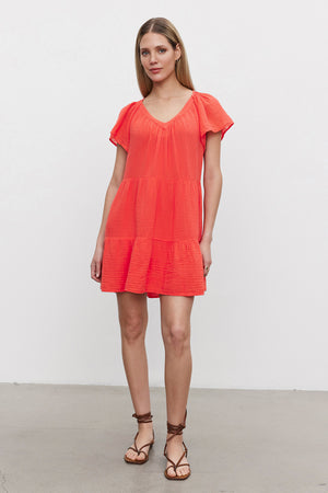 A person stands wearing the ELEANOR COTTON GAUZE DRESS by Velvet by Graham & Spencer, featuring a short-sleeve design in red with a ruffled bottom and frilled flutter sleeves, paired with brown strappy sandals against a plain white background.