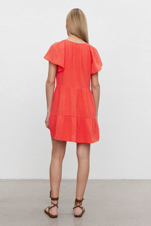 A woman with long blonde hair stands facing away from the camera, wearing the ELEANOR COTTON GAUZE DRESS by Velvet by Graham & Spencer, an orange short-sleeve tiered dress with frilled flutter sleeves, paired with brown strappy sandals.