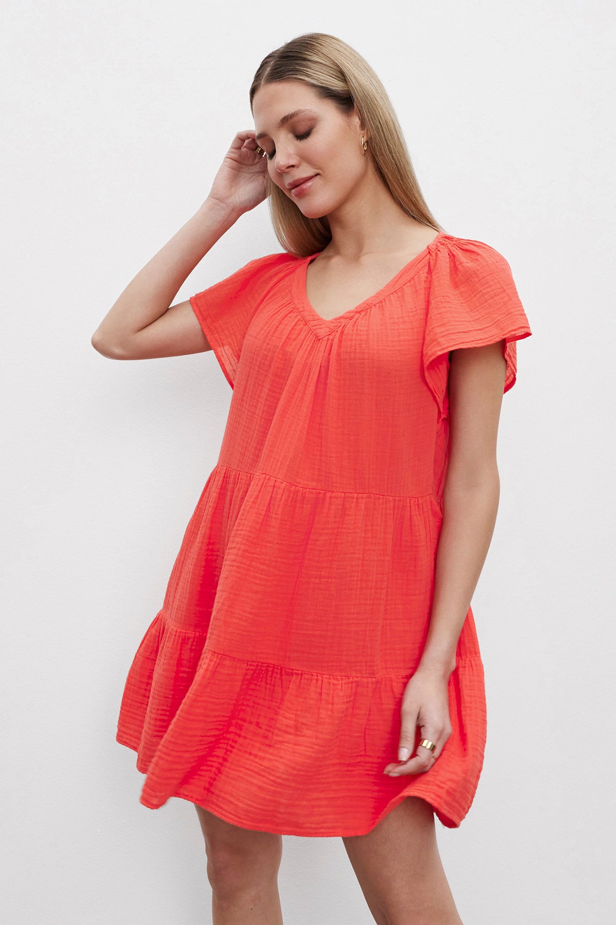A woman with light brown hair is wearing the ELEANOR COTTON GAUZE DRESS by Velvet by Graham & Spencer, a bright pink short-sleeved dress featuring a V-neck and tiered silhouette. She is looking down, one hand touching her ear, against a plain white background. The airy cotton gauze fabric adds an extra touch of grace to her ensemble.-37618635800769