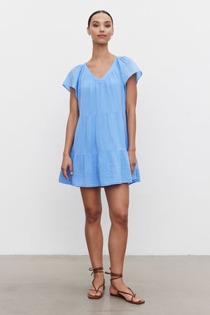A person stands against a plain white background wearing the Velvet by Graham & Spencer ELEANOR COTTON GAUZE DRESS in light blue, featuring a tiered silhouette, paired with brown strappy sandals.