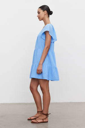 A woman wearing the ELEANOR COTTON GAUZE DRESS by Velvet by Graham & Spencer stands sideways to the camera. The light blue, short-sleeve dress features a tiered silhouette and falls gracefully as she stands on a bare concrete floor against a plain white wall. She pairs the airy cotton gauze dress with brown strappy sandals and has her hair pulled back.
