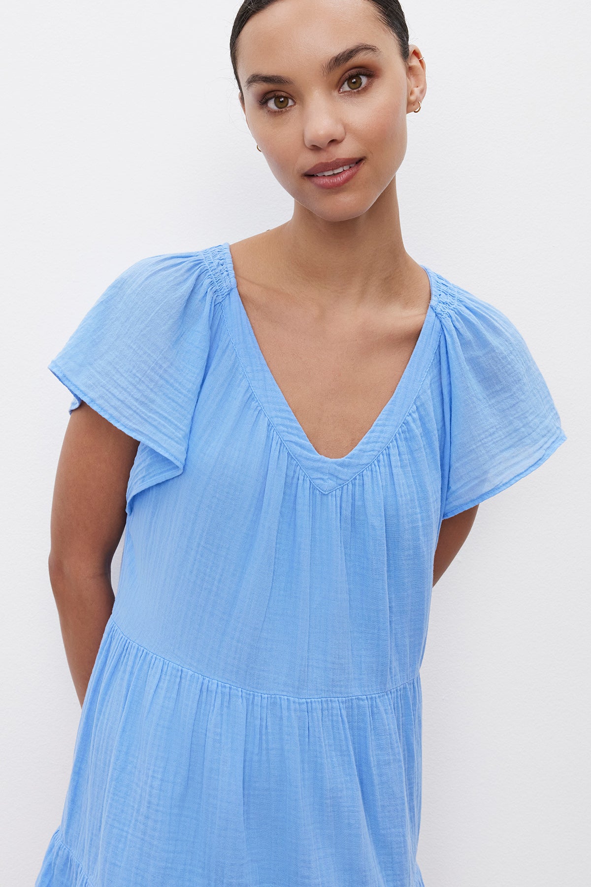 A woman wearing the ELEANOR COTTON GAUZE DRESS by Velvet by Graham & Spencer, featuring frilled flutter sleeves and a tiered silhouette in light blue, stands against a white background, looking at the camera.