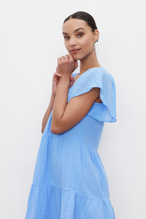 A person stands against a white background, wearing the ELEANOR COTTON GAUZE DRESS by Velvet by Graham & Spencer in light blue short sleeves, looking at the camera with hands near the face.