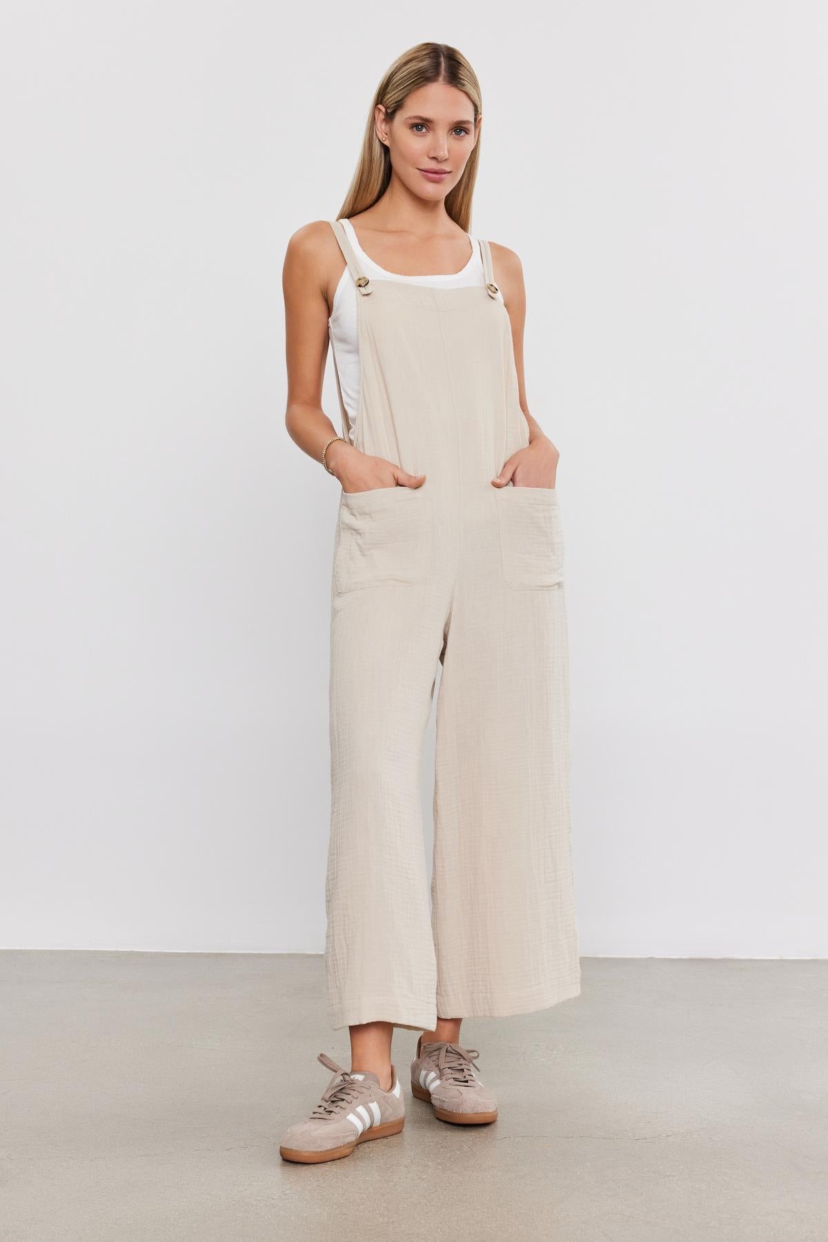 A woman stands in a studio, wearing a white tank top and Velvet by Graham & Spencer beige cotton gauze jumpsuit with patch pockets, paired with brown sneakers, looking directly at the camera.
