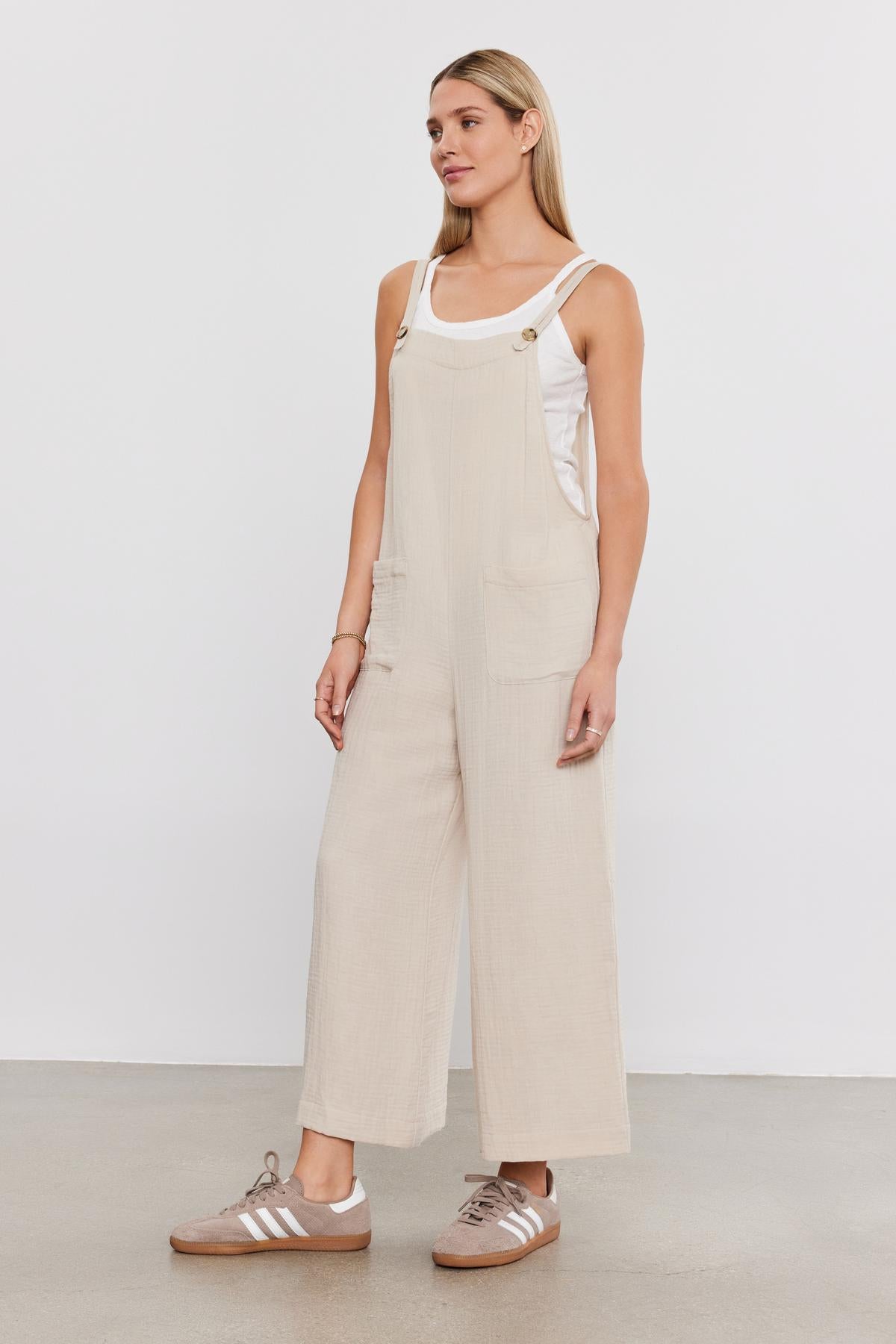 Woman standing in a studio, wearing a Velvet by Graham & Spencer EVERLEE COTTON GAUZE JUMPSUIT with patch pockets and brown sneakers, looking to her left with a neutral expression.