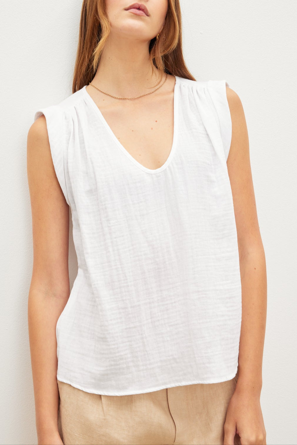 A person with long hair is wearing the Velvet by Graham & Spencer JAYLA COTTON GAUZE TANK TOP, a sleeveless white V-neck top with subtle shirred detailing, and beige pants, standing against a plain white background.-37618902302913