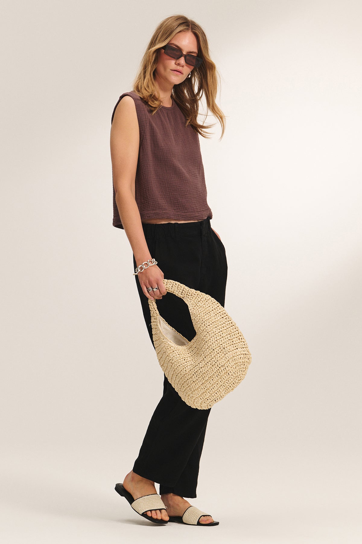 A woman wearing sunglasses, a sleeveless brown top, black pants, and sandals holds the SHELLY BAG by Velvet by Graham & Spencer while standing against a plain background.-39292578791617