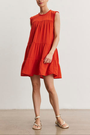 A woman stands indoors wearing the SUSIE COTTON GAUZE DRESS by Velvet by Graham & Spencer and sandals. She has her hair tied back and is looking at the camera. The background wall is plain white.