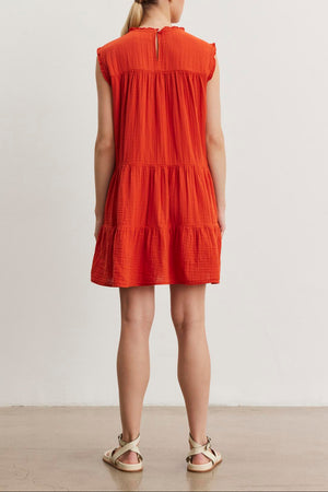 A person with long, blonde hair and wearing a short-sleeved, orange SUSIE COTTON GAUZE DRESS from Velvet by Graham & Spencer stands facing away from the camera. The dress has a tiered design and the individual is wearing white sandals.