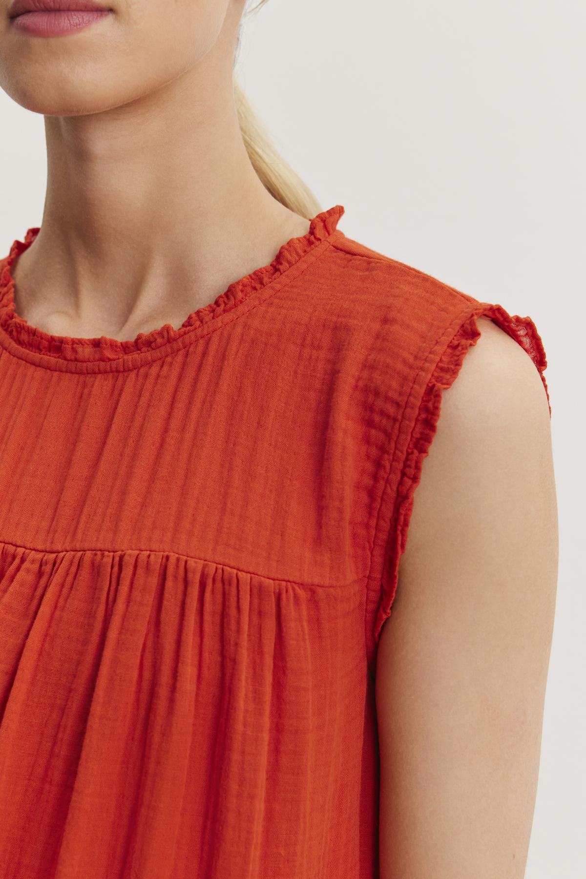 A close-up of a person wearing a sleeveless, ruffled neckline orange SUSIE COTTON GAUZE DRESS by Velvet by Graham & Spencer. The person has light skin and blonde hair tied back.-37074513232065