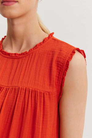 A close-up of a person wearing a sleeveless, ruffled neckline orange SUSIE COTTON GAUZE DRESS by Velvet by Graham & Spencer. The person has light skin and blonde hair tied back.