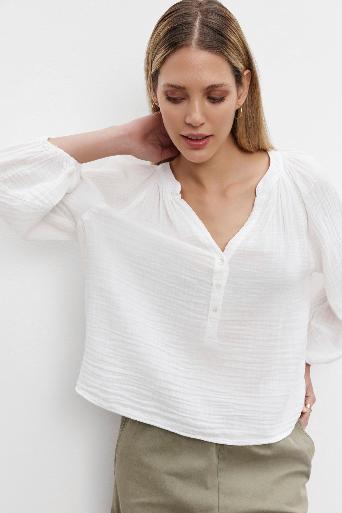 A woman with long hair wears the VIVI COTTON GAUZE TOP by Velvet by Graham & Spencer. The white, airy blouse features a V-neck and button details. She poses with one hand behind her head, highlighting the relaxed fit and lightweight fabric of the top.