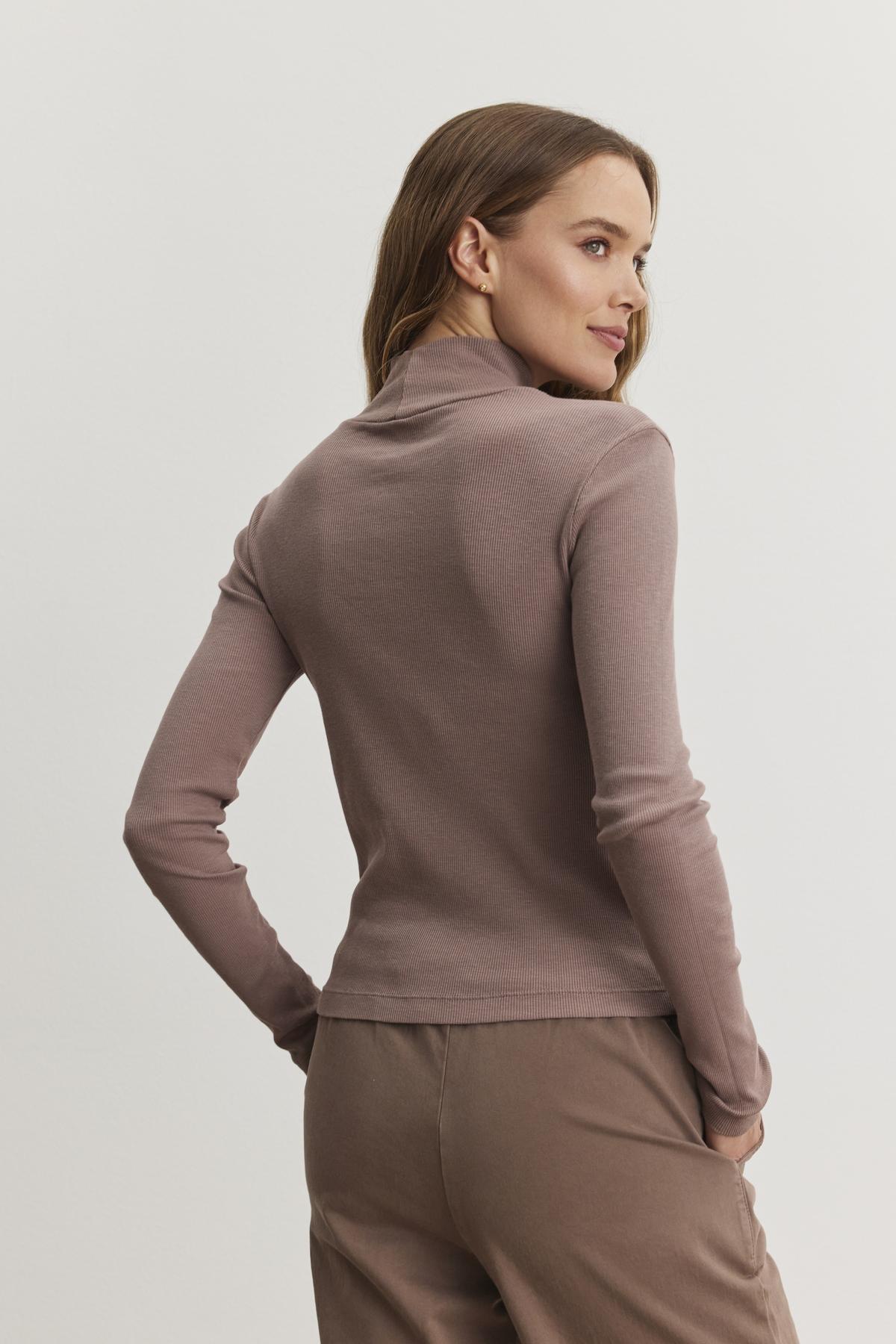 A person is wearing the ADRIAN TEE by Velvet by Graham & Spencer, a mauve ribbed cotton slub turtleneck with brown pants, slightly turned to the side against a plain background.-38483543818433