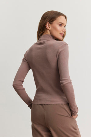 A person is wearing the ADRIAN TEE by Velvet by Graham & Spencer, a mauve ribbed cotton slub turtleneck with brown pants, slightly turned to the side against a plain background.