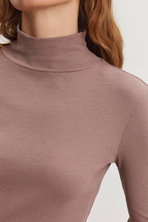 A person is wearing an ADRIAN TEE from Velvet by Graham & Spencer, which is a brown ribbed cotton slub turtleneck; their shoulder-length brown hair contrasts against a plain background.