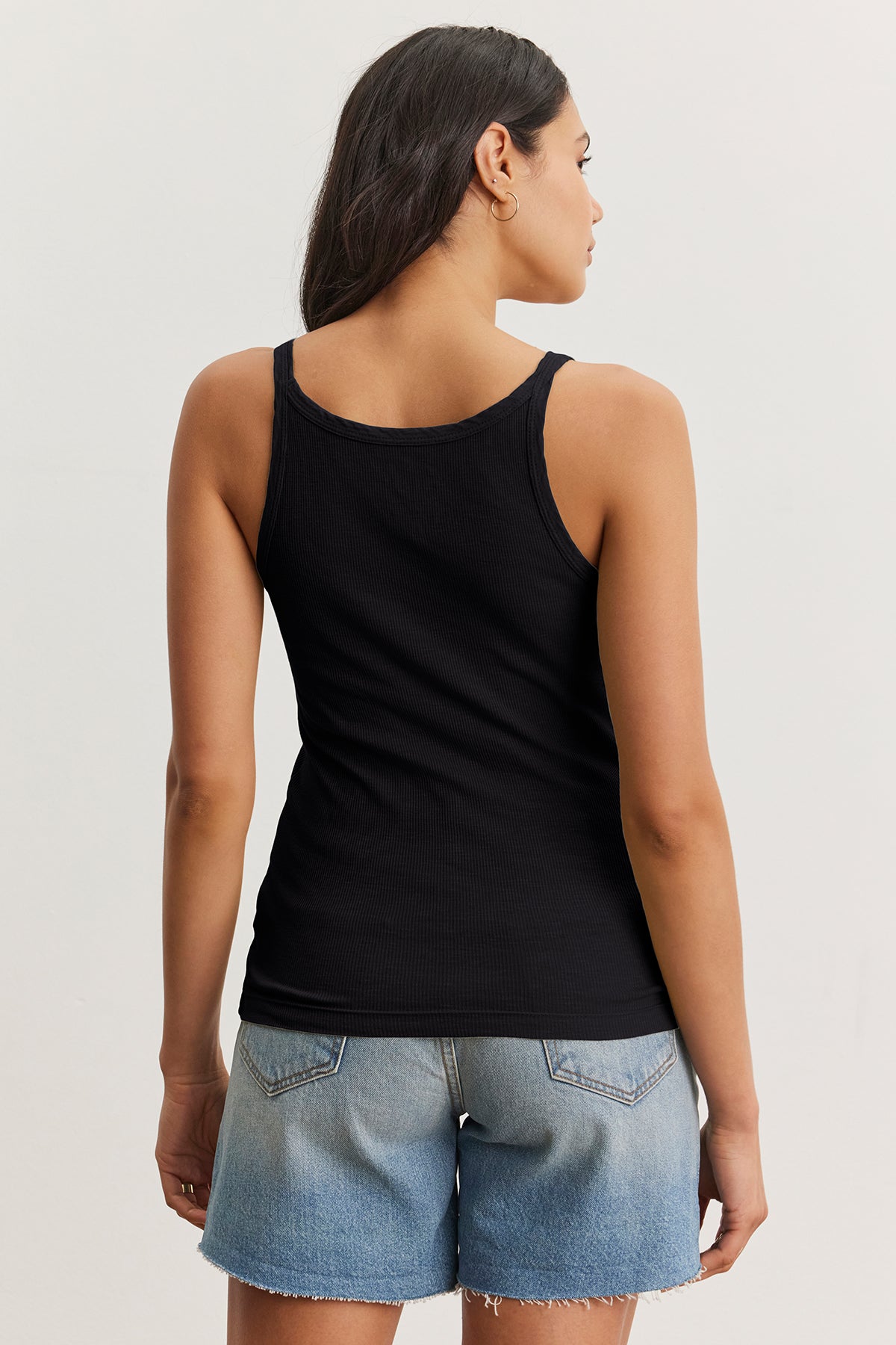 A woman with long hair stands with her back to the camera, wearing a Velvet by Graham & Spencer ALIZA RIBBED COTTON LAYERING TANK in black and denim shorts.-38807335403713