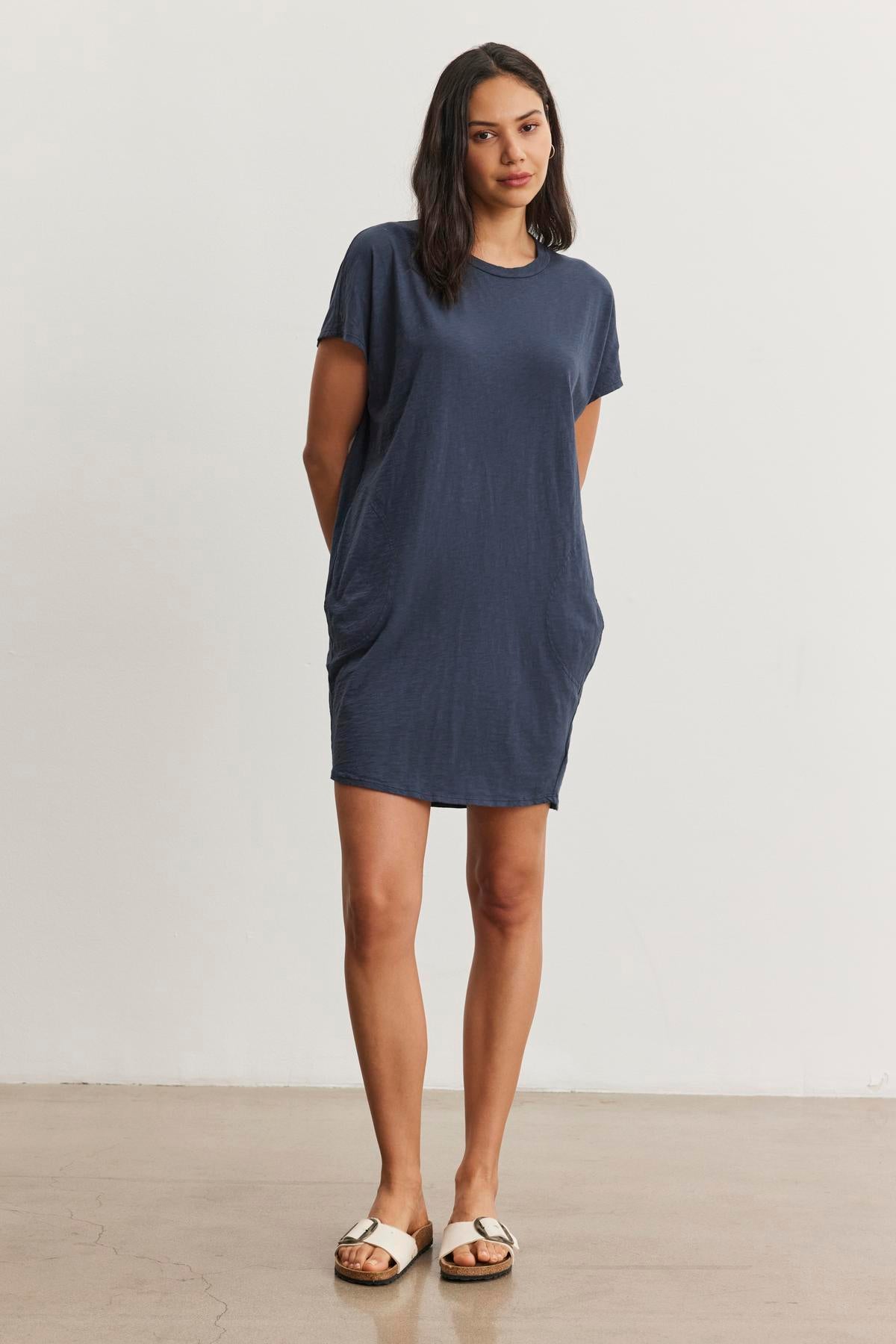 A person with long hair stands wearing a relaxed fit, short-sleeve blue ATHENA DRESS by Velvet by Graham & Spencer with hands in pockets and white sandals, against a plain white background.