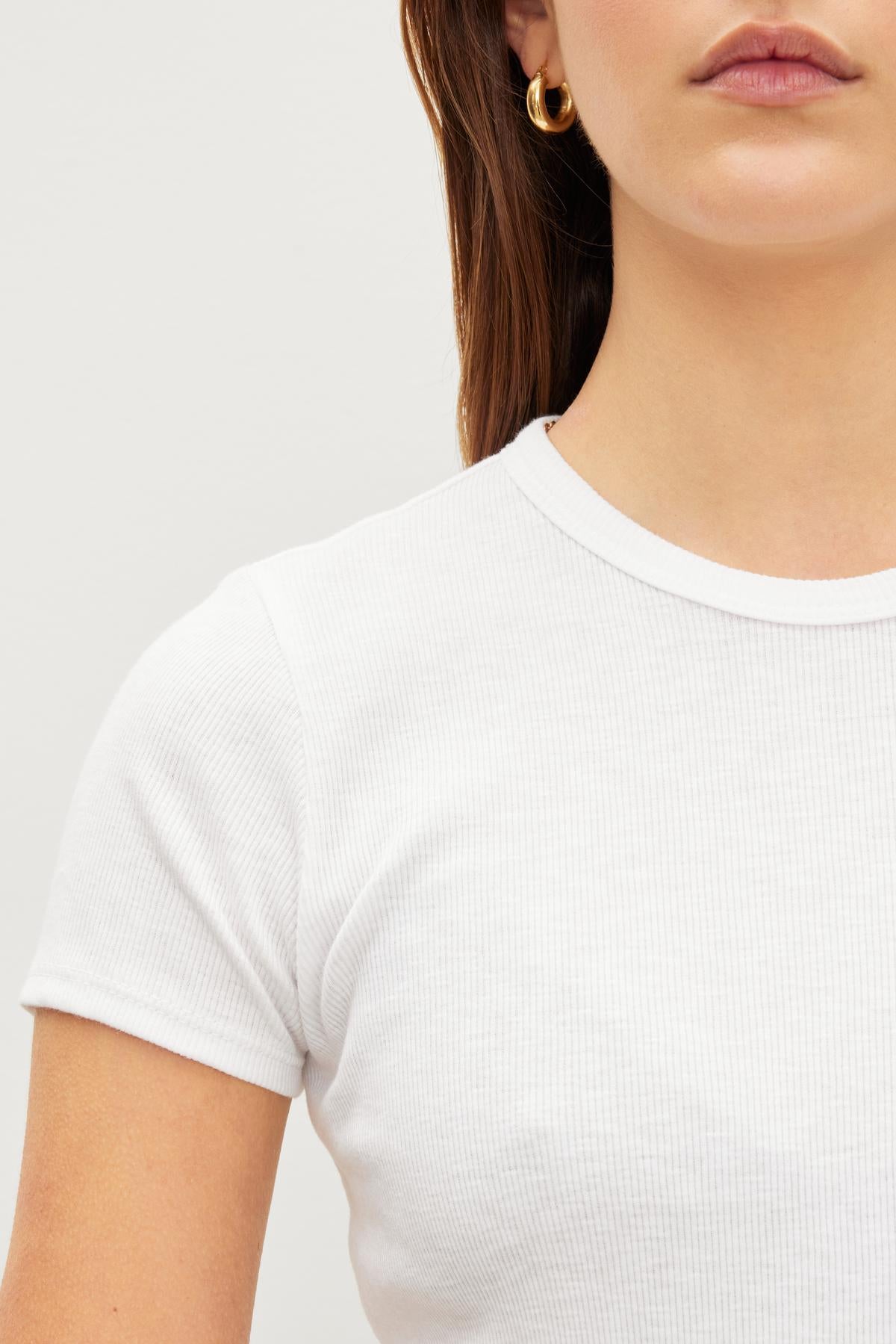 A woman is shown from the shoulders up against a plain background, wearing Velvet by Graham & Spencer's BRENNY TEE—the white ribbed cotton knit T-shirt with a crew neckline—and gold hoop earrings.-35955654688961
