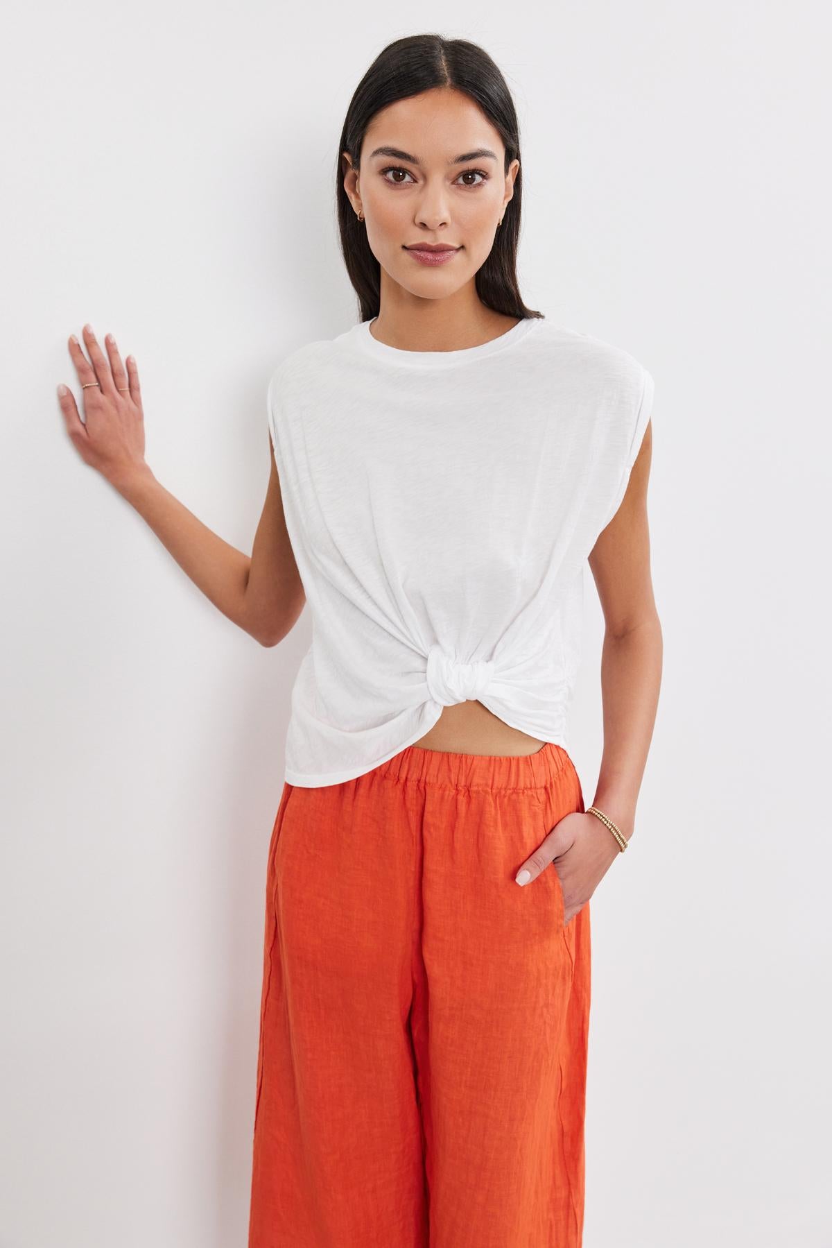 A woman in a Velvet by Graham & Spencer CALLISTA TEE with twist knot detail and orange pants standing against a white background, with one hand on her hip and the other raised slightly.-36909996409025