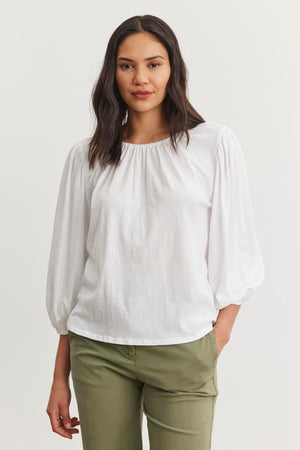 A person stands against a plain background, wearing the CARENA TEE by Velvet by Graham & Spencer, made from lightweight cotton slub with puff sleeves, paired with olive green pants.