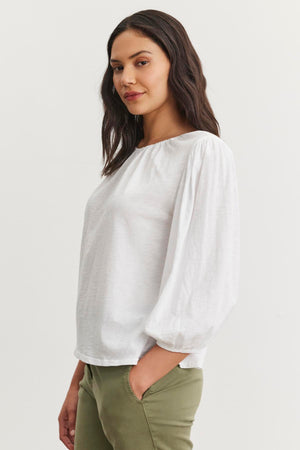 Against a plain background, a person wears the CARENA TEE—a lightweight cotton slub blouse with an elastic neckline and puffed sleeves by Velvet by Graham & Spencer—paired with olive green pants.