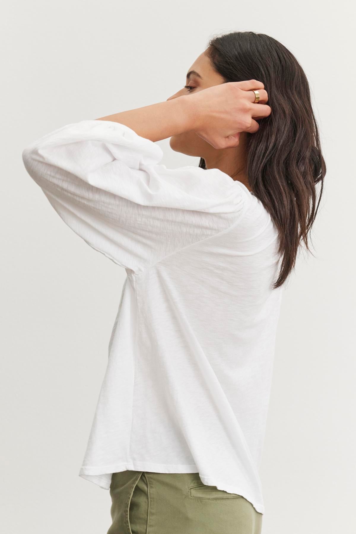 A long-haired person in a white CARENA TEE by Velvet by Graham & Spencer adjusts their hair. The tee's lightweight cotton slub fabric ripples slightly as they move, wearing green pants against a neutral backdrop.-38499462021313