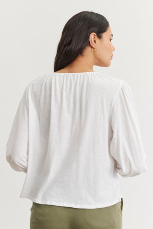 The person with long hair is seen from the back wearing a Velvet by Graham & Spencer CARENA TEE, a white long-sleeve top with an elastic neckline and puff sleeves, paired with green pants. The outfit is crafted from lightweight cotton slub against a plain background.